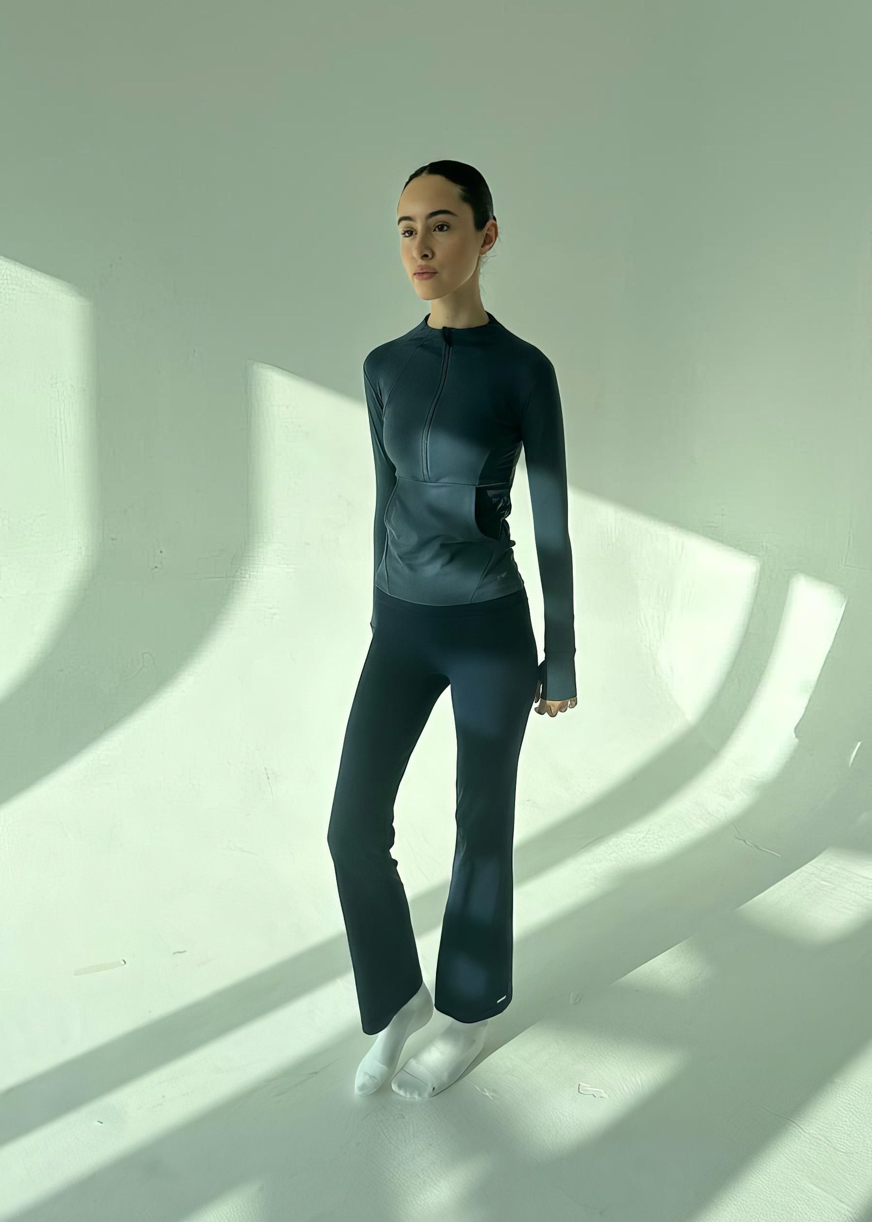 Person wearing a dark outfit standing in a minimalistic setting with light casting shadows.