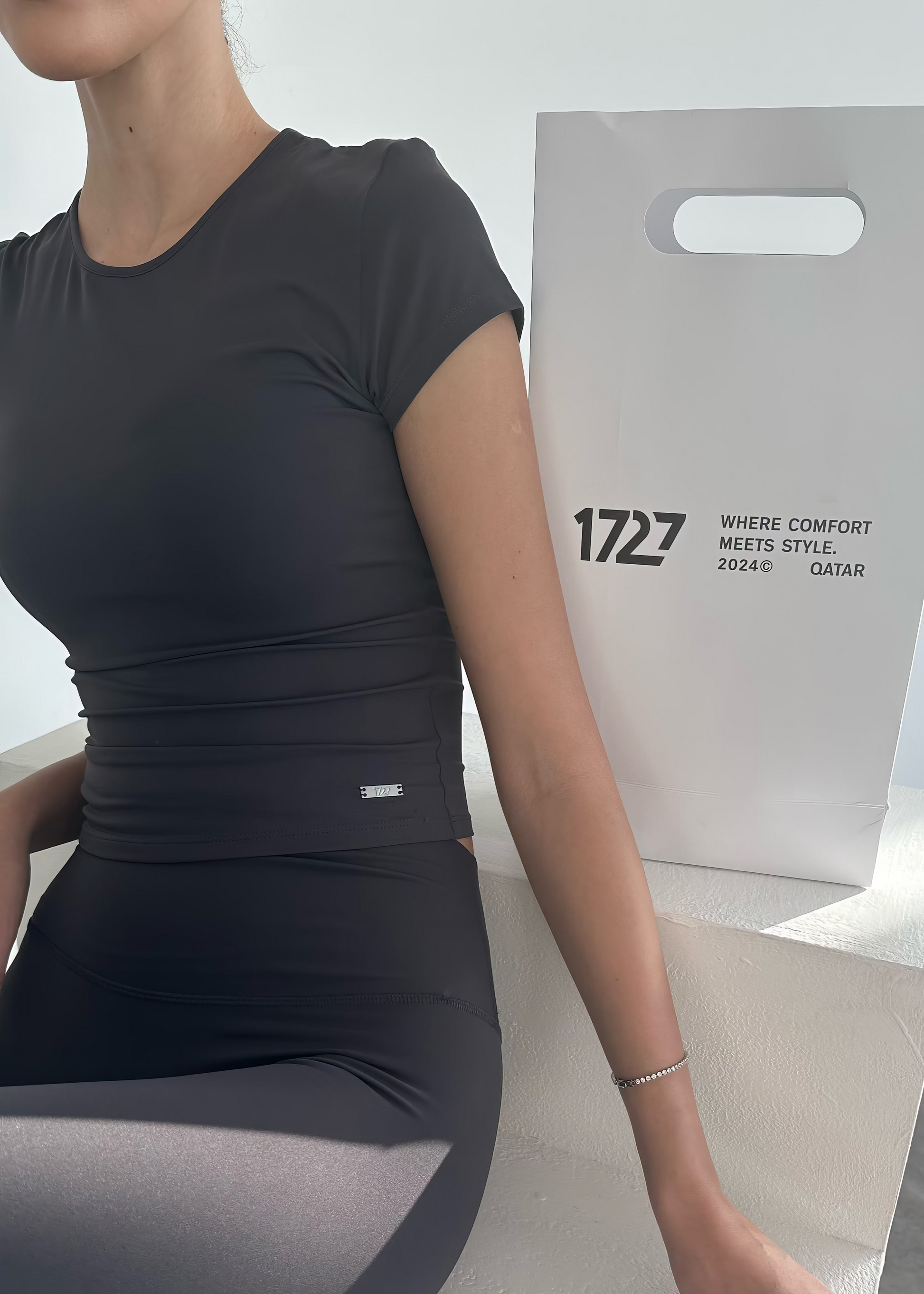 Person wearing a black athletic outfit sitting on a white surface with a branded box in the background.