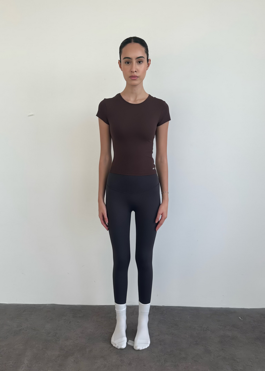 Person wearing a brown t-shirt and black leggings standing against a white wall.