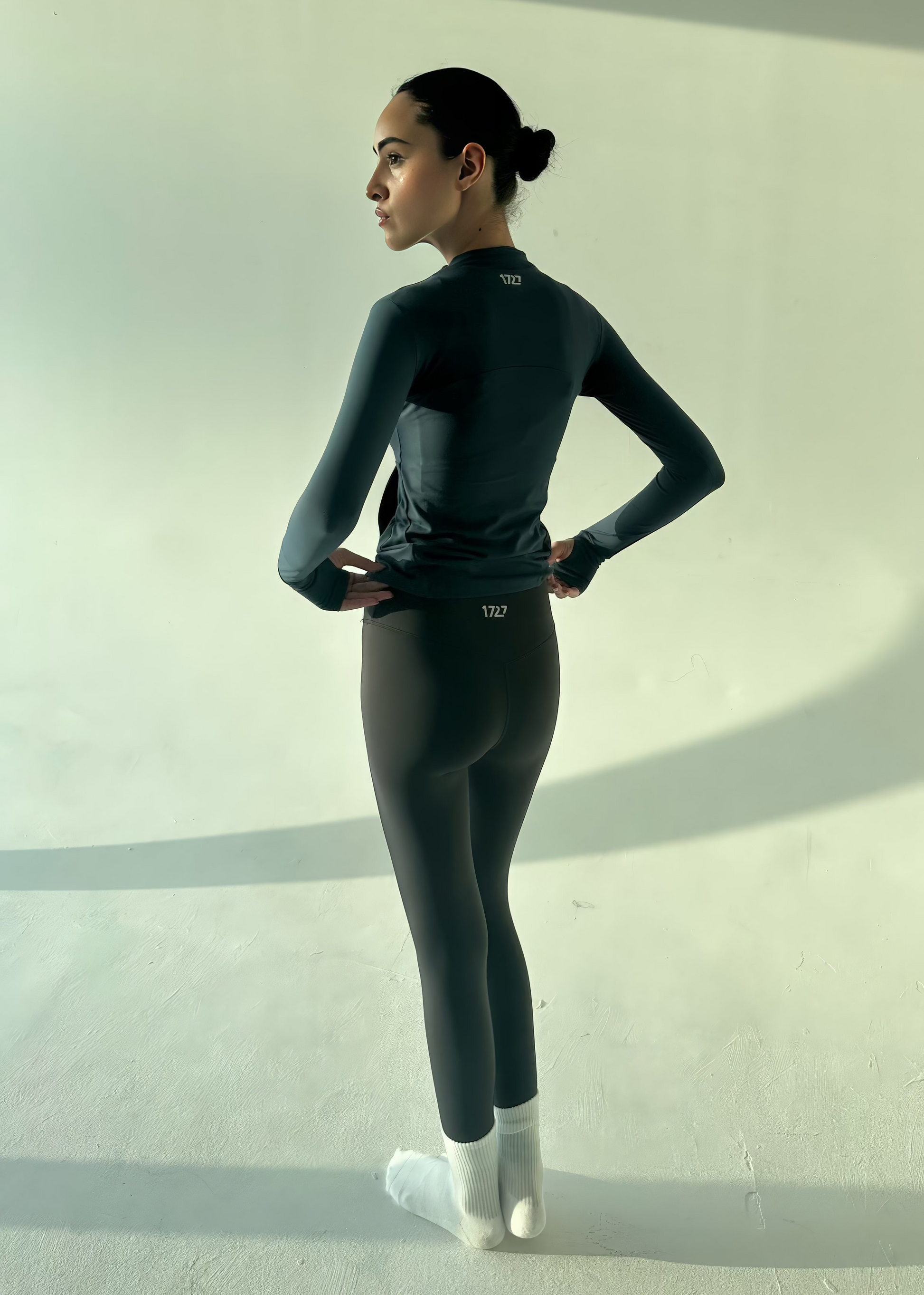 Person wearing a black long-sleeve top and leggings standing against a white wall.