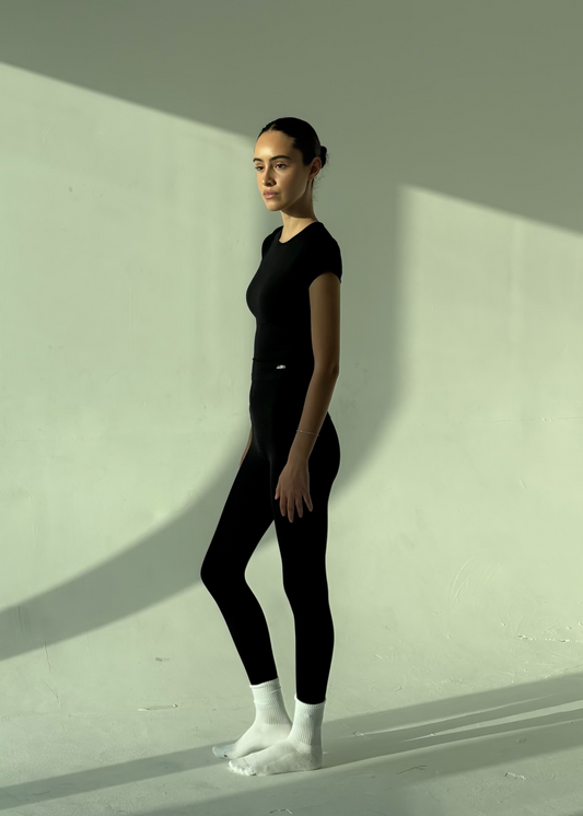 Person wearing a black outfit standing against a white wall with shadows.